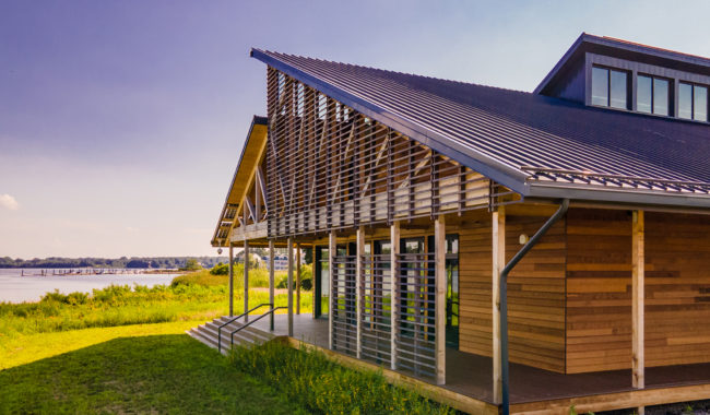 A modern wooden building with a slanted metal roof sits by a grassy area near a body of water, bathed in sunlight under a clear, blue sky.