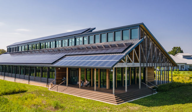 A modern, two-story building with large windows and angled roofs covered in solar panels, surrounded by green grass and vegetation under a clear blue sky.