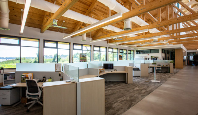 Modern open office with high wooden ceilings, large windows letting in natural light, carpeted floors, light-colored cubicles, and ergonomic chairs. Several desks have computers and office supplies.