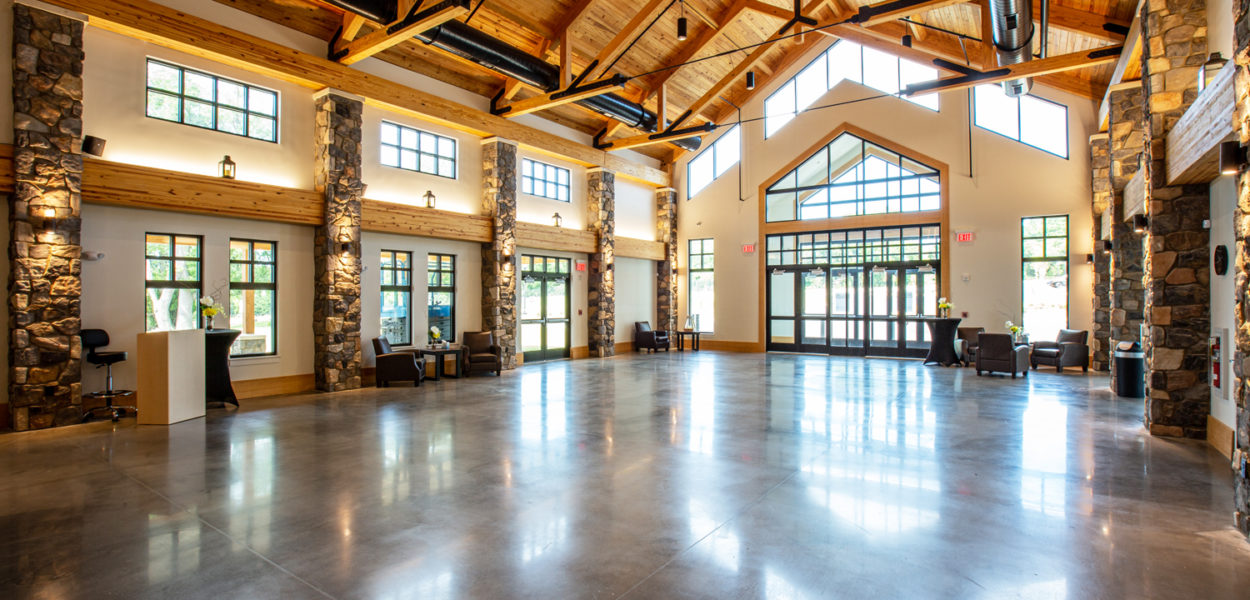 Spacious hall with polished concrete floors, exposed wooden beams, stone pillars, large windows, and glass doors letting in natural light. Several chairs and a desk are positioned along the walls.