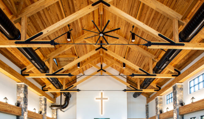 Interior view of a church with exposed wooden beams, large black ceiling fans, stone pillars, and a glowing cross mounted on the front wall above two screens. Natural light streams through side windows.