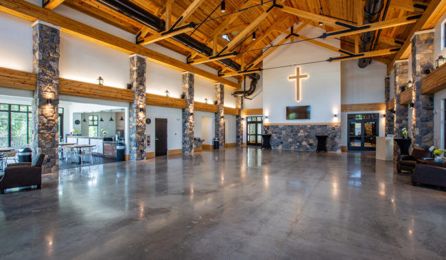 Large, modern church lobby with high wooden ceilings, exposed beams, stone pillars, polished concrete floor, a prominent cross on the wall, couches, tables, and large windows letting in natural light.