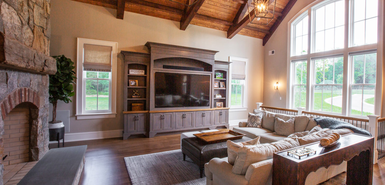 A spacious living room with vaulted wooden ceiling, large arched windows, a stone fireplace, a gray sectional sofa, a wooden entertainment center with a TV, and soft natural lighting.