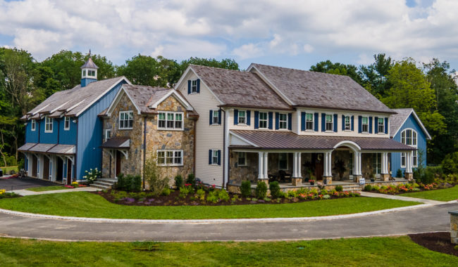 A large, elegant house with stone and blue siding, a covered front porch, and a three-car garage, surrounded by landscaped gardens and a curved driveway, with trees and a cloudy sky in the background.