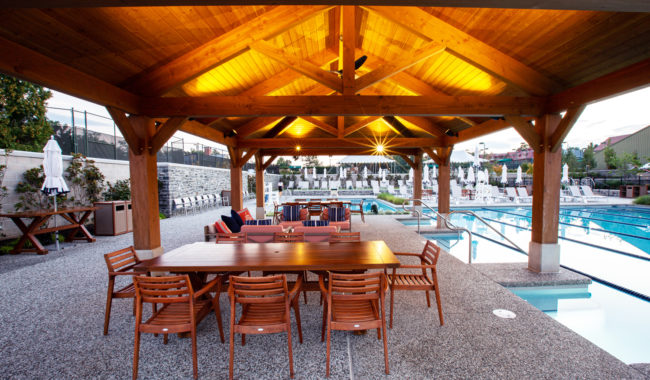A wooden gazebo with a lit ceiling covers a wooden dining table and chairs beside a pool. Lounge chairs and umbrellas line the pool deck, creating a relaxing outdoor setting.