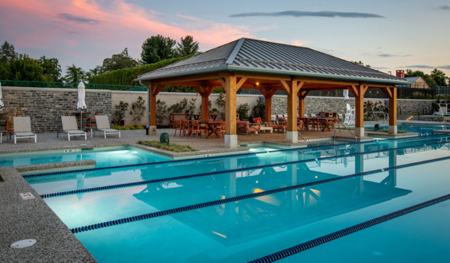 Outdoor swimming pool with lap lanes, lounge chairs, and a wooden pavilion with tables and chairs underneath. The pool area is surrounded by a stone wall and trees at sunset, with a colorful sky overhead.