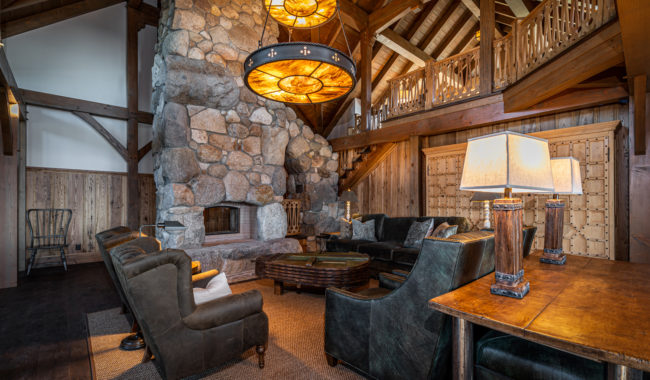 A spacious rustic living room with high wooden beams, a large stone fireplace, cozy dark armchairs, a wooden table with lamps, and two large circular chandeliers hanging from the ceiling.
