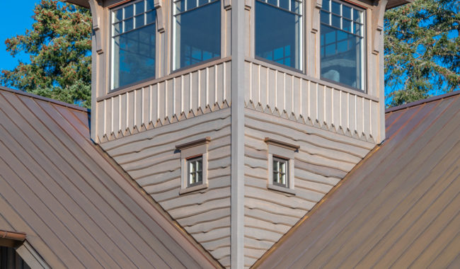 A wooden building with a steep brown metal roof features a prominent tower with multiple windows. The structure has decorative trim and is set against a clear blue sky and green trees.