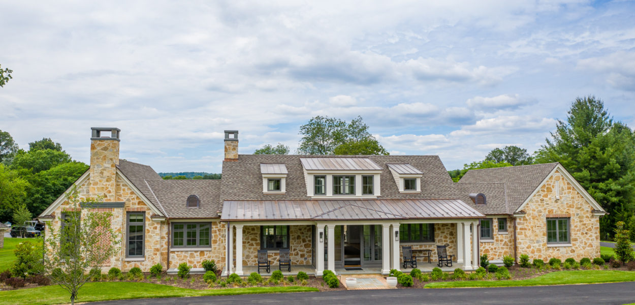 A large stone house with a metal roof, multiple chimneys, and dormer windows sits surrounded by green lawns, shrubs, and trees under a partly cloudy sky. A curved driveway leads to the front porch.