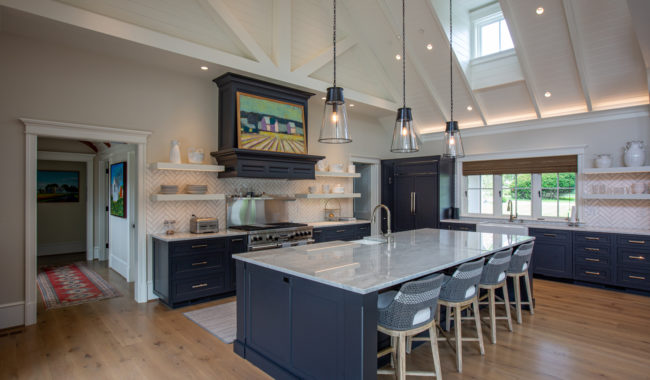 A spacious, modern kitchen with a large marble island, four woven barstools, dark blue cabinets, open shelves with dishes, pendant lights, and vaulted white ceilings with exposed beams. A range hood displays a colorful painting.