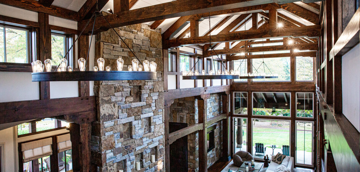 A spacious living room with high ceilings, exposed wooden beams, large windows, and a stone fireplace. The room features modern furniture, a chandelier with exposed bulbs, and natural light streaming in.