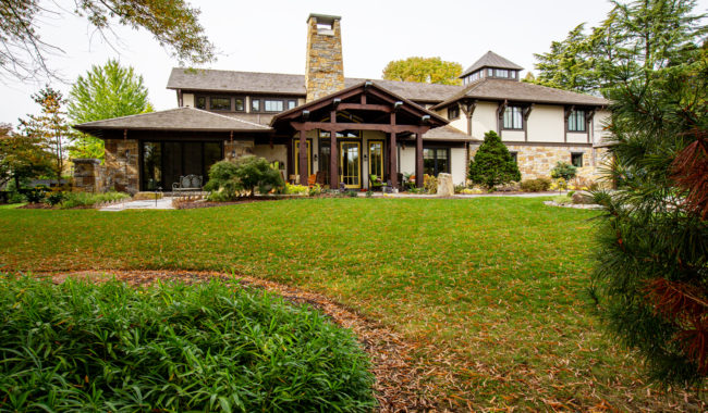 A large, modern house with stone and wood accents, tall windows, and a central stone chimney, surrounded by landscaped gardens and green lawn under an overcast sky.