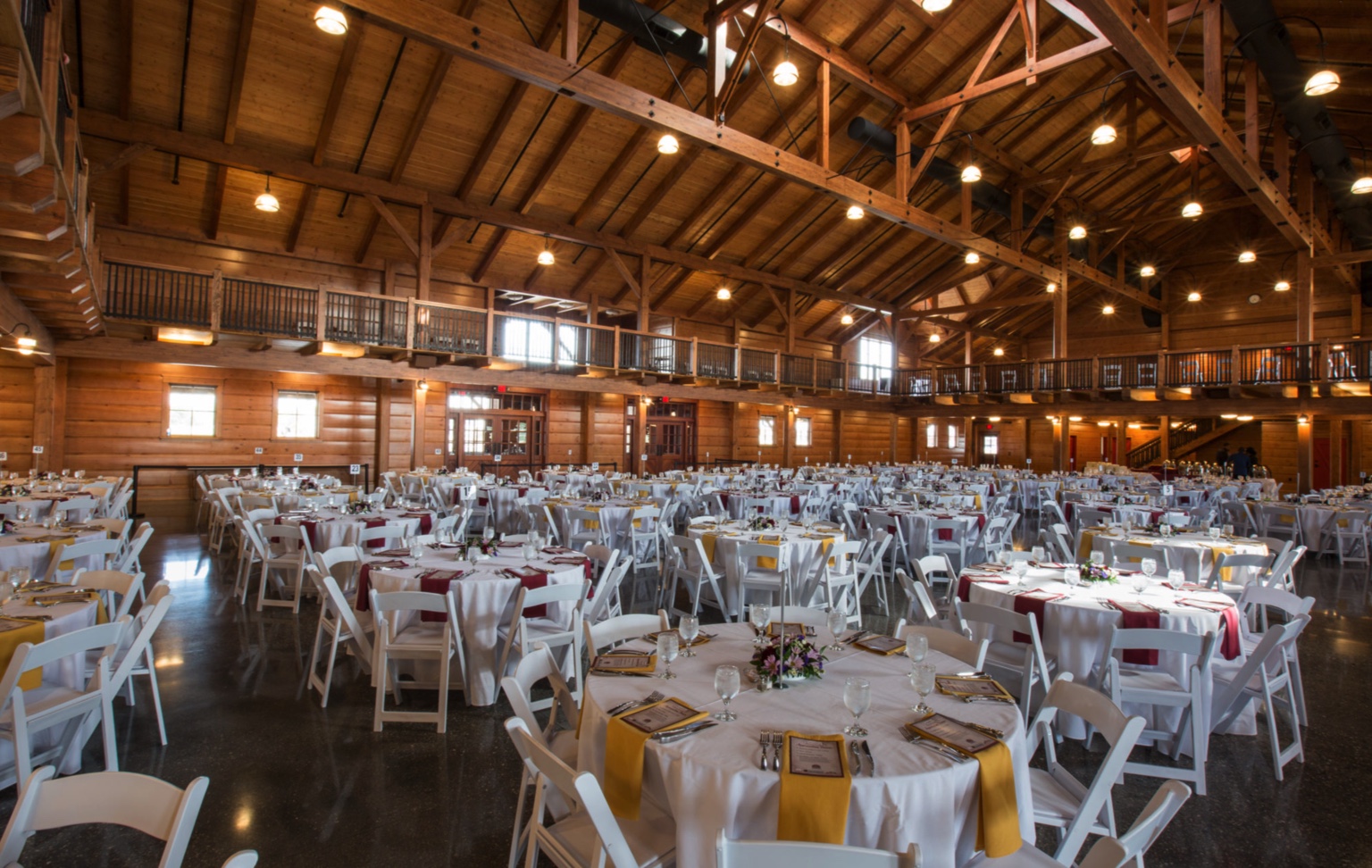 A spacious, rustic banquet hall with timber frame beams and high ceilings, featured in our project gallery, filled with round tables set with white chairs, tableware, and centerpieces—perfectly ready for a formal event.