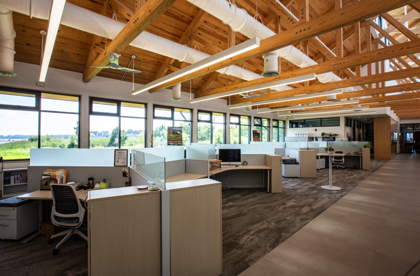A modern, open office space with light wood beams, large windows, and cubicles features in this timber frame project gallery. The area is bright and spacious, filled with natural light and offering views of lush greenery outside.