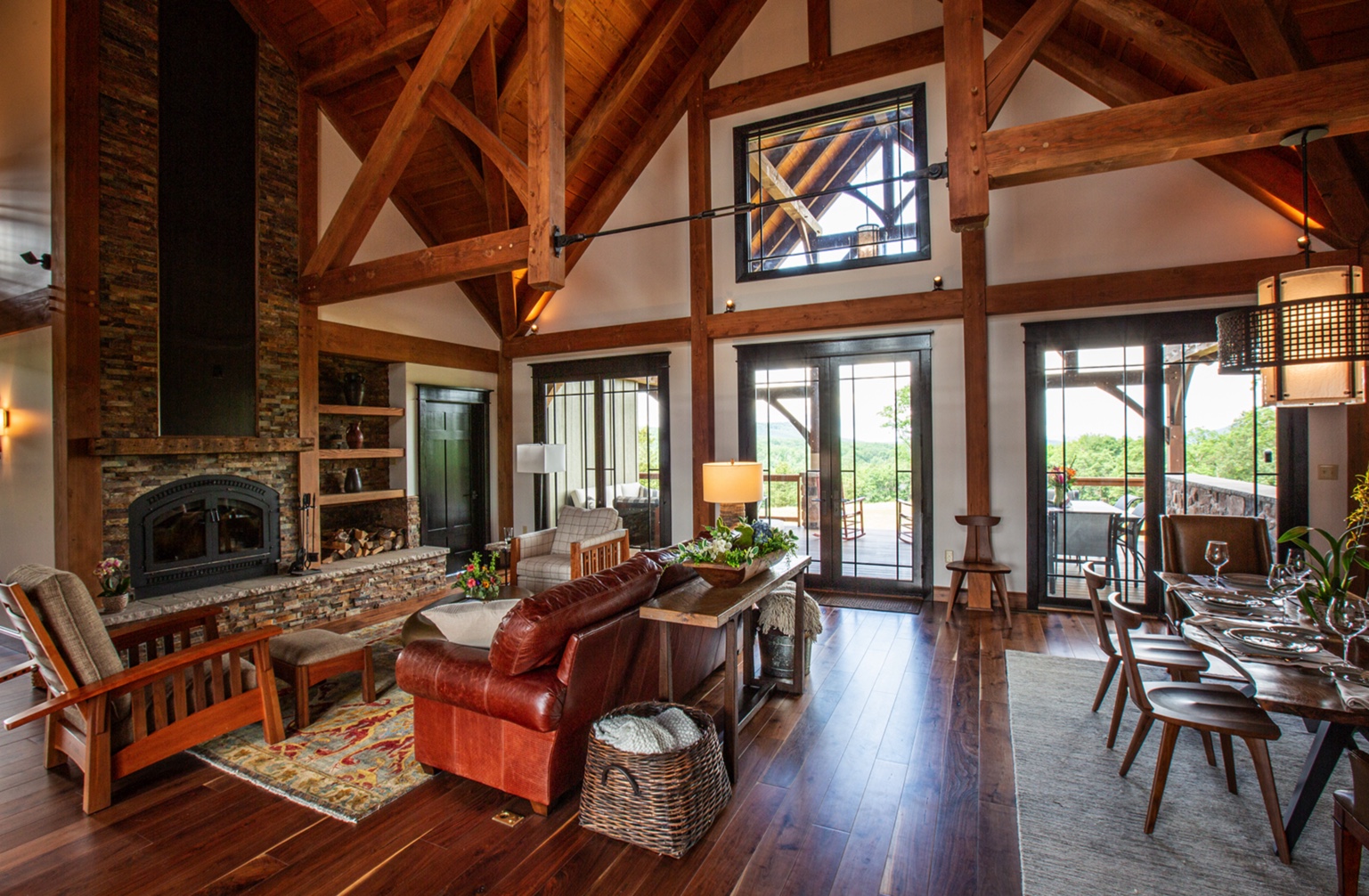 A spacious, rustic living and dining area with exposed wooden beams, a stone fireplace, and large windows—this timber frame project gallery showcases leather and wooden furniture, plus glass doors opening to an outdoor patio with lush greenery views.