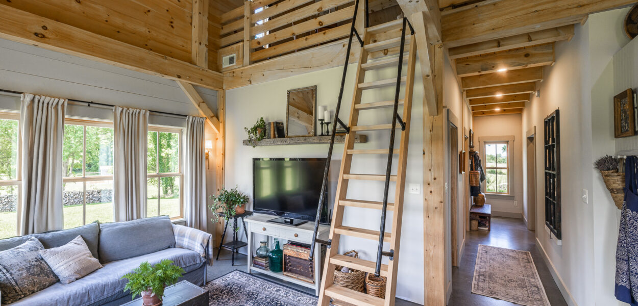 A cozy, modern tiny house interior features wood beams, a loft accessible by a ladder, a living area with a couch and TV, large windows with curtains, and a hallway leading to the back of the home.