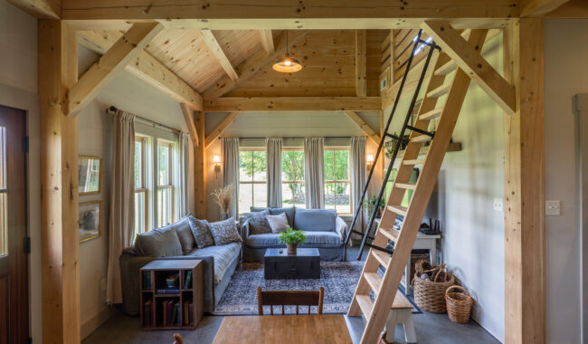 A cozy living room with exposed wooden beams, a loft with a ladder, gray sectional sofas, a rug, large windows with curtains, a coffee table, bookshelves, and baskets on the floor. Natural light fills the space.