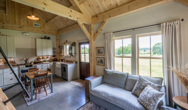 Cozy, rustic living space with wooden beams, a gray sofa, kitchen area with white cabinets, dining table, and large windows letting in natural light and offering a view of green fields outside.