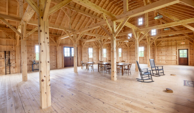 Spacious interior of a wooden barn with exposed beams, wooden floors, large windows, scattered tables and chairs, and two rocking chairs, creating a rustic, airy atmosphere.