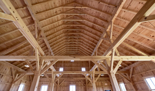 Interior view of a large wooden barn with exposed beams, high vaulted ceiling, and multiple windows allowing natural light to fill the spacious, unfinished room.