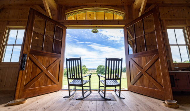 Two wooden rocking chairs sit on a rug inside a wooden cabin with wide open double doors, facing a scenic view of green fields and a blue sky with clouds.