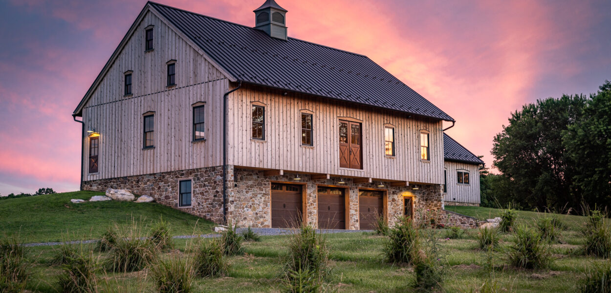 A rustic barn with stone and wood siding stands on a grassy hill at sunset, illuminated by soft exterior lights. The sky glows with pink and purple tones as the sun sets behind the building.