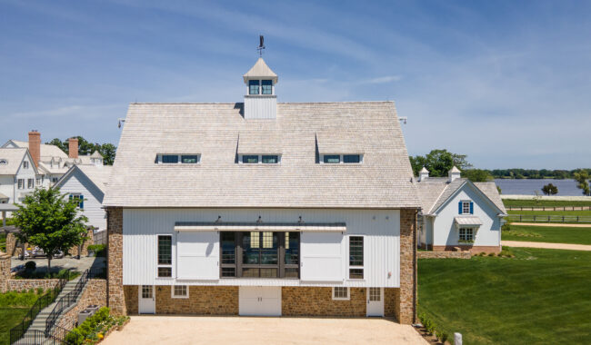 A large, white barn with stone lower walls, multiple windows, and a cupola sits on well-manicured green lawns, with a winding path, additional buildings, and a river in the background under a blue sky.
