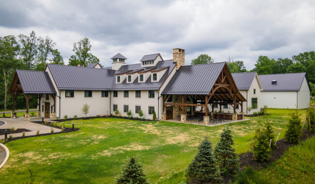 A large, modern farmhouse-style building with white siding, a dark metal roof, and exposed wooden beams, surrounded by green lawns, small trees, and a cloudy sky.