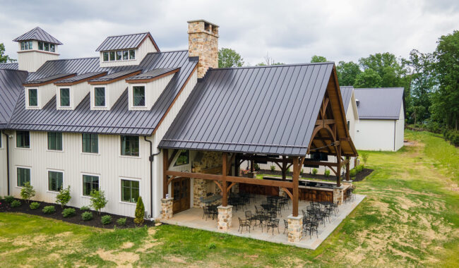 A large modern farmhouse with white siding and a dark metal roof features a covered outdoor patio with tables and chairs. The patio has wooden beams and stone pillars, surrounded by green grass and landscaping.