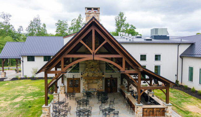 A large modern farmhouse with white siding, a dark metal roof, and a spacious outdoor patio featuring wooden beams, metal chairs, tables, and a stone fireplace under a covered pergola. Trees and cloudy sky are in the background.