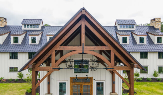 A close-up view of a large, modern barn-style building with a metal roof and wooden beams over the entrance, featuring multiple windows and landscaped greenery.