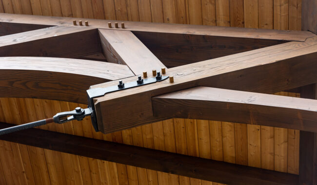 Close-up of a wooden roof truss with metal fasteners and bolts, showing detailed craftsmanship and joinery against a wooden ceiling background.