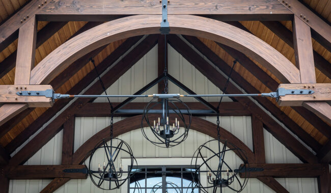 Wooden ceiling beams and arches support metal light fixtures with exposed bulbs, set against a cream-colored paneled wall. The architecture has a rustic, industrial style.