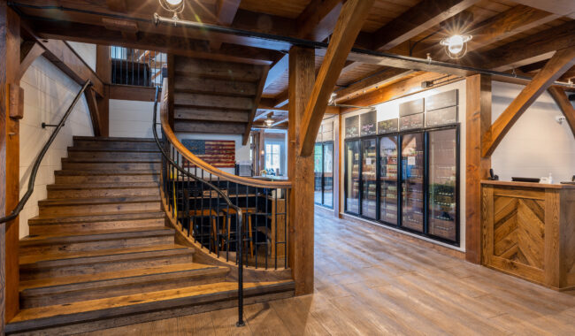 A spacious interior with wooden beams, a curved wooden staircase with black railing, large glass refrigerator units along one wall, and a wood-accented counter; the space has warm lighting and a rustic, modern design.