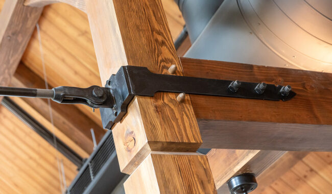 Close-up of a large black metal bracket bolted to a wooden beam, supporting the timber structure of a ceiling with visible wood paneling and exposed ducts in the background.