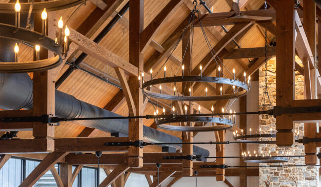 Exposed wooden beams and circular metal chandeliers with small lit bulbs hang from a high, vaulted ceiling inside a rustic, modern building with stone and wooden accents.