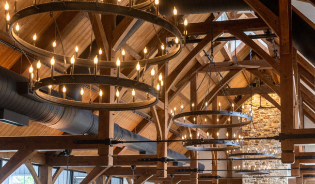 Large circular chandeliers with lit bulbs hang from exposed wooden beams in a high, vaulted ceiling. The space features stone walls and industrial black air ducts, creating a rustic, elegant atmosphere.