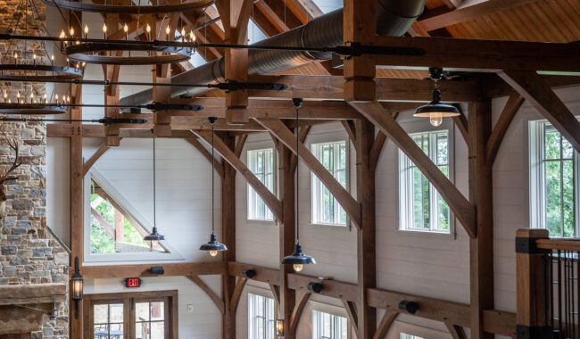 Interior of a rustic building with exposed wooden beams, large windows letting in natural light, stone fireplace, and modern hanging light fixtures suspended from the ceiling.