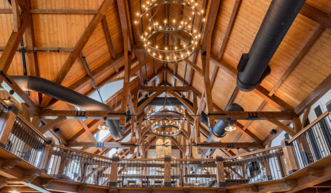 Interior of a building with exposed wooden beams, vaulted ceilings, black metal railings, round chandeliers with many lights, and large black ventilation ducts, creating a rustic yet modern atmosphere.