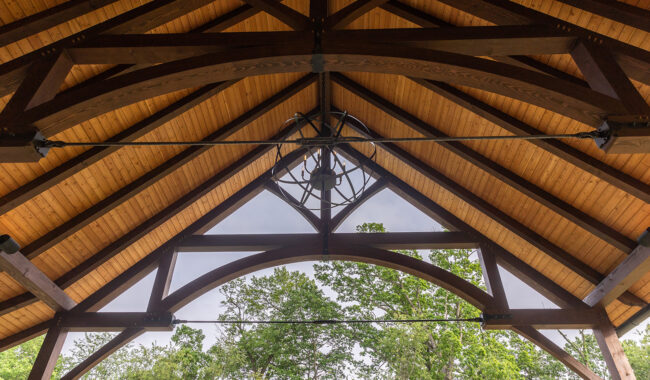 View of a wooden pavilion ceiling with exposed beams and a central chandelier. Trees and a cloudy sky are visible through the open side of the structure.