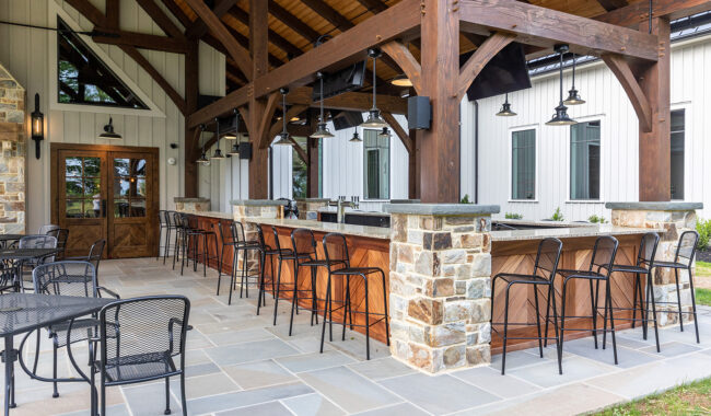 An outdoor bar area with a wooden roof, stone pillars, high bar stools, and metal patio tables and chairs, next to a modern building with white siding and large windows.