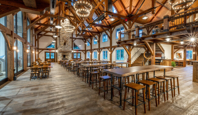 Spacious dining hall with wooden beams, high ceilings, long tables, and chairs. Large stone fireplace at the end, tall windows let in natural light, and modern chandeliers hang overhead. Rustic yet elegant atmosphere.
