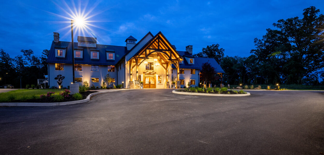 A large, elegant building with peaked roofs and wooden beams is lit warmly at dusk, surrounded by trees and landscaped gardens, with a circular driveway in front and a lamppost casting a bright light.