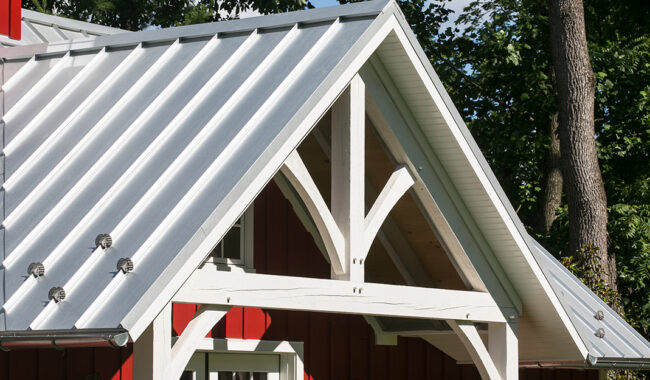 Close-up of a house with a white metal roof and decorative white wooden gable trim, contrasting with red vertical siding, surrounded by green trees in the background.