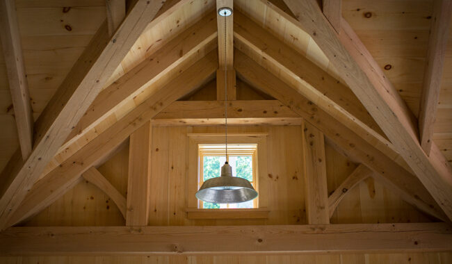 A metal pendant light hangs from the ceiling inside a wooden cabin, with exposed beams and a small window letting in daylight above the light fixture.