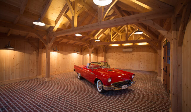 A classic red convertible car with whitewall tires is parked inside a spacious wooden garage with exposed beams, brick flooring, and warm overhead lighting.