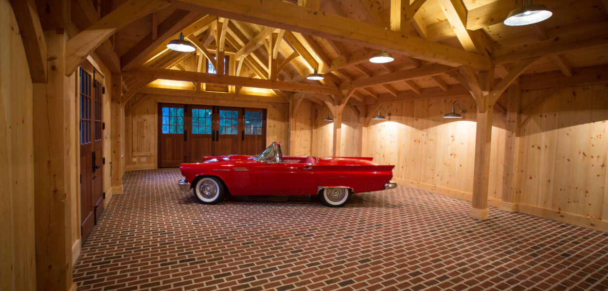A classic red convertible car is parked inside a spacious wooden garage with exposed beams, brick flooring, and large windows at the far end.