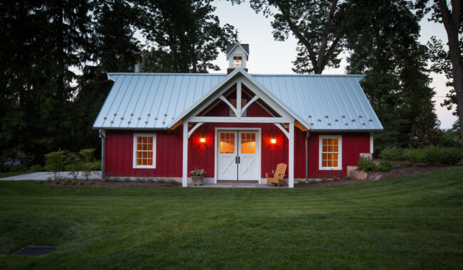 A small red barn with a white roof and trim, glowing exterior lights, double white doors, and two yellow chairs on a covered porch, surrounded by green grass and tall trees at dusk.