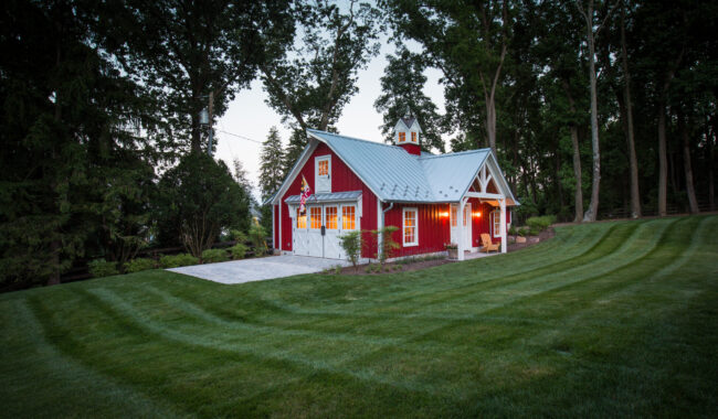 A small red house with white trim and a metal roof sits among tall trees, surrounded by a well-manicured lawn with striped mowing patterns. Warm lights glow from the windows and front porch.