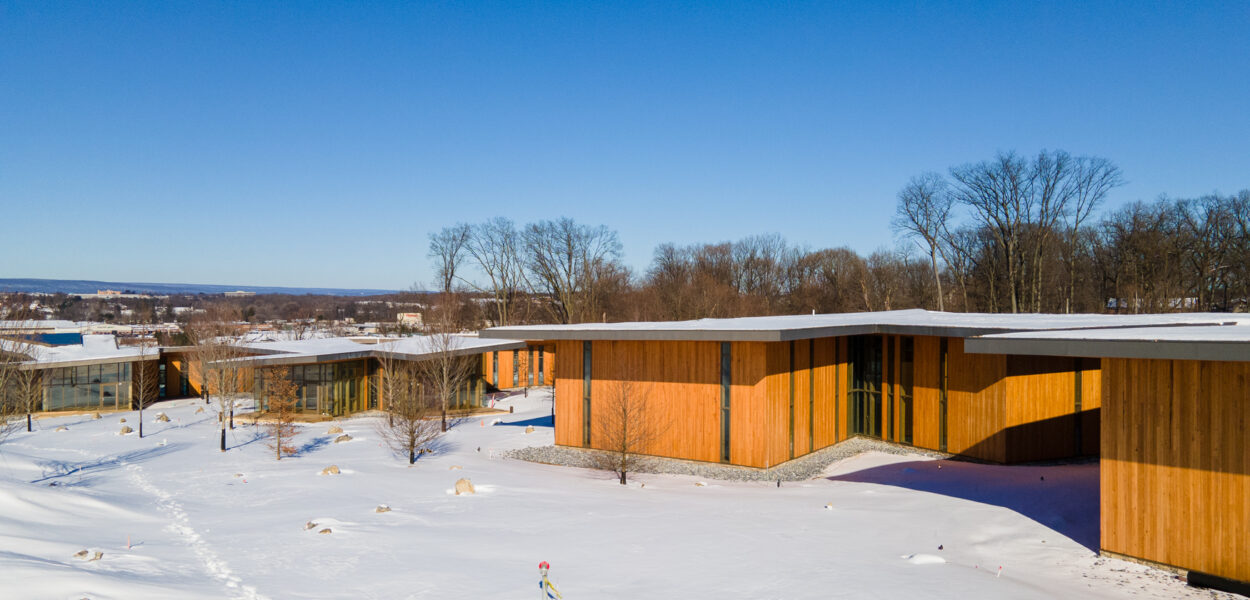 Modern wooden buildings with large windows are set among leafless trees and snow-covered ground on a clear, sunny day with a bright blue sky.